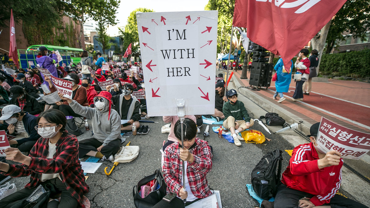 Protesters sit in the street, with one holding a large sign reading "I'm with her" with arrows pointing outward in all directions